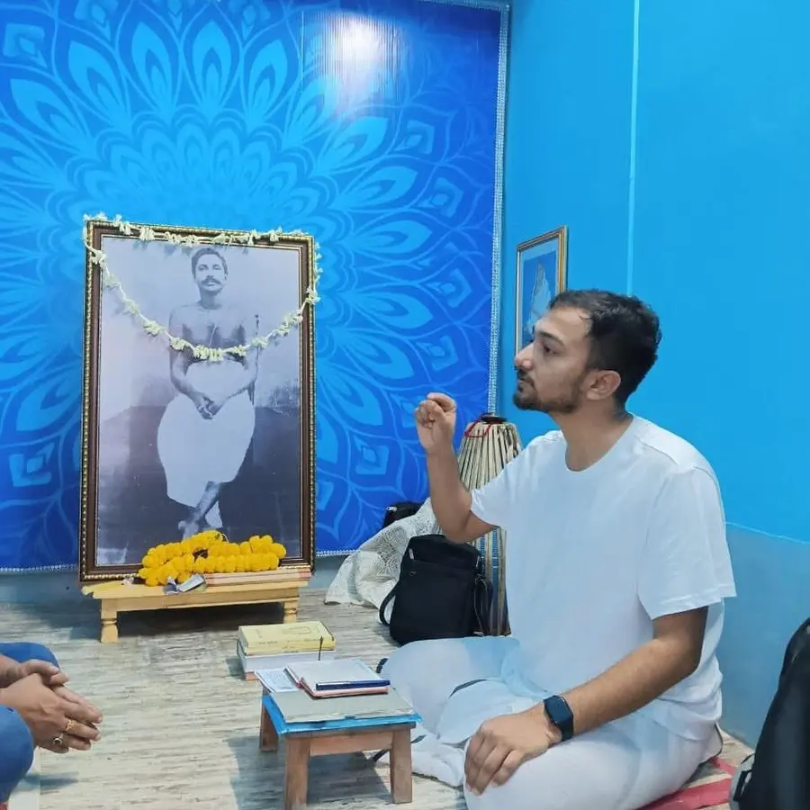 Souvik Ghosh seated on the floor in white, speaking, beside a garlanded portrait of Sree Sree Thakur Anukulchandra.