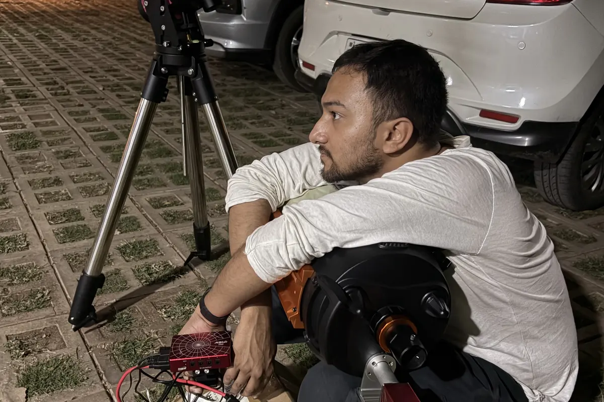 Souvik Ghosh sitting outdoors at night beside the tripod, holding the controller, with the tube beside him.