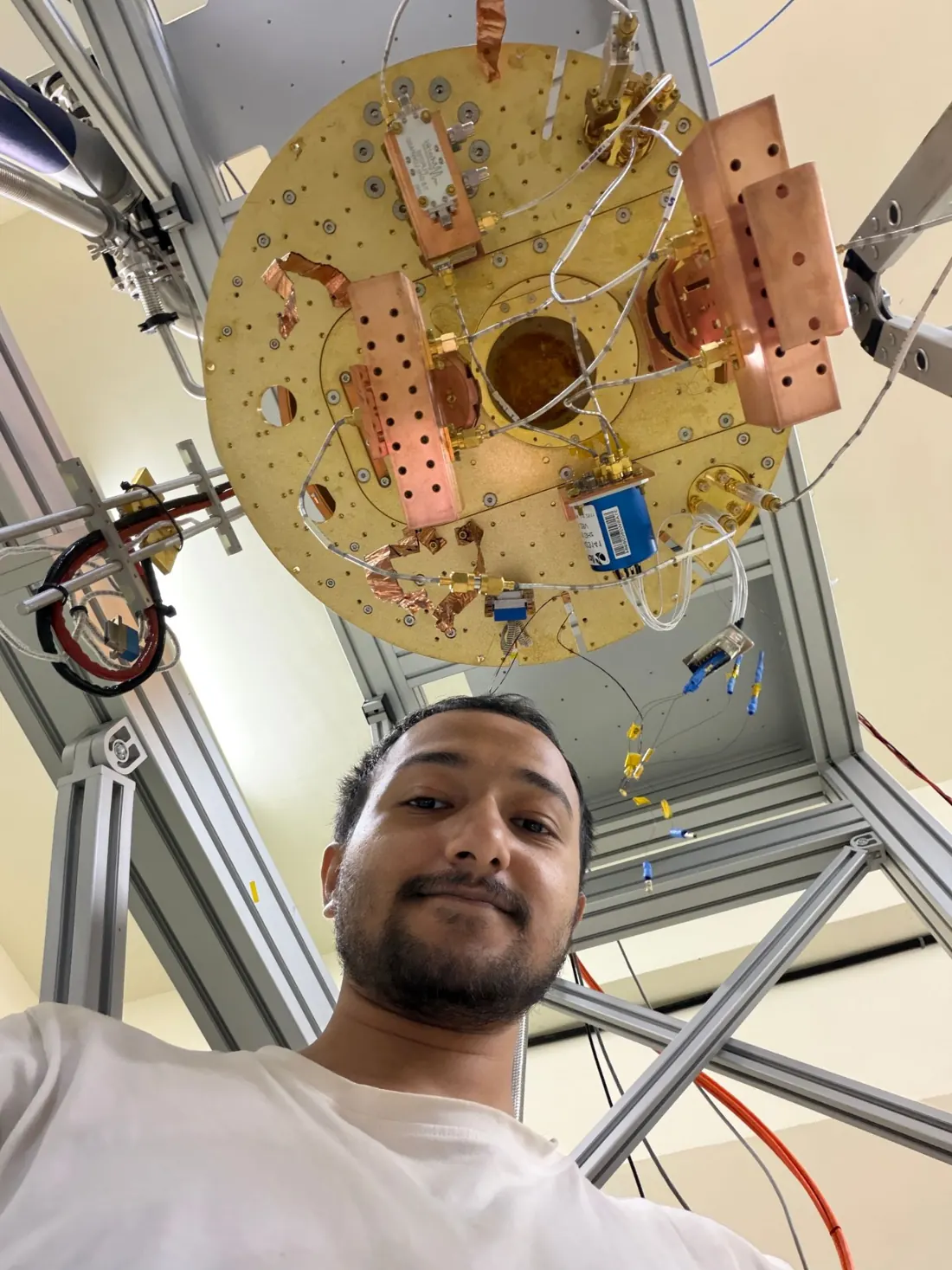 Souvik Ghosh looking up at the camera from under the lowest plate of a dilution refrigerator, which carries copper brackets, microwave cables and a blue switch.