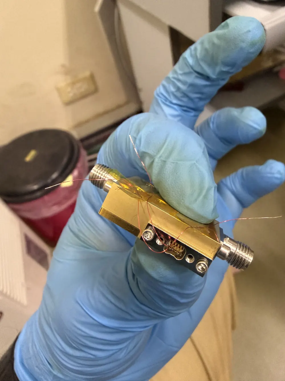 A gloved hand holding a small gold-plated sample holder with two threaded connectors and loose fine wires.