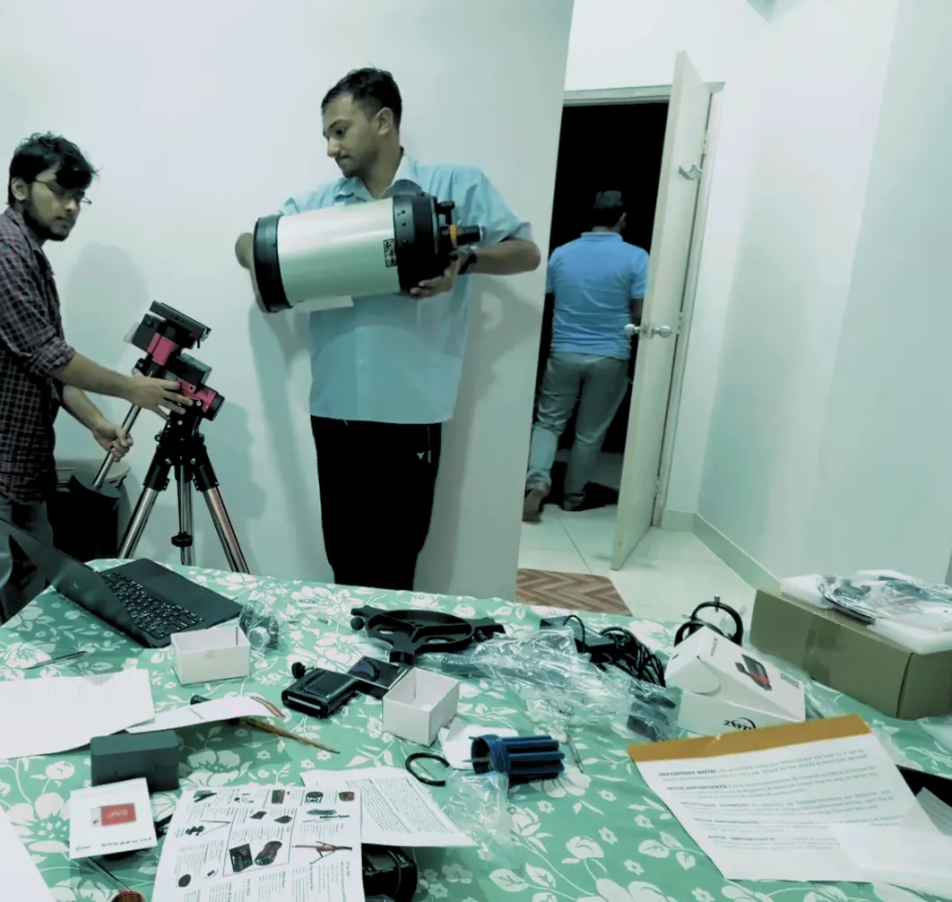 Two people in a room, one steadying the mount on its tripod, the other holding the optical tube, with manuals and parts on a table.
