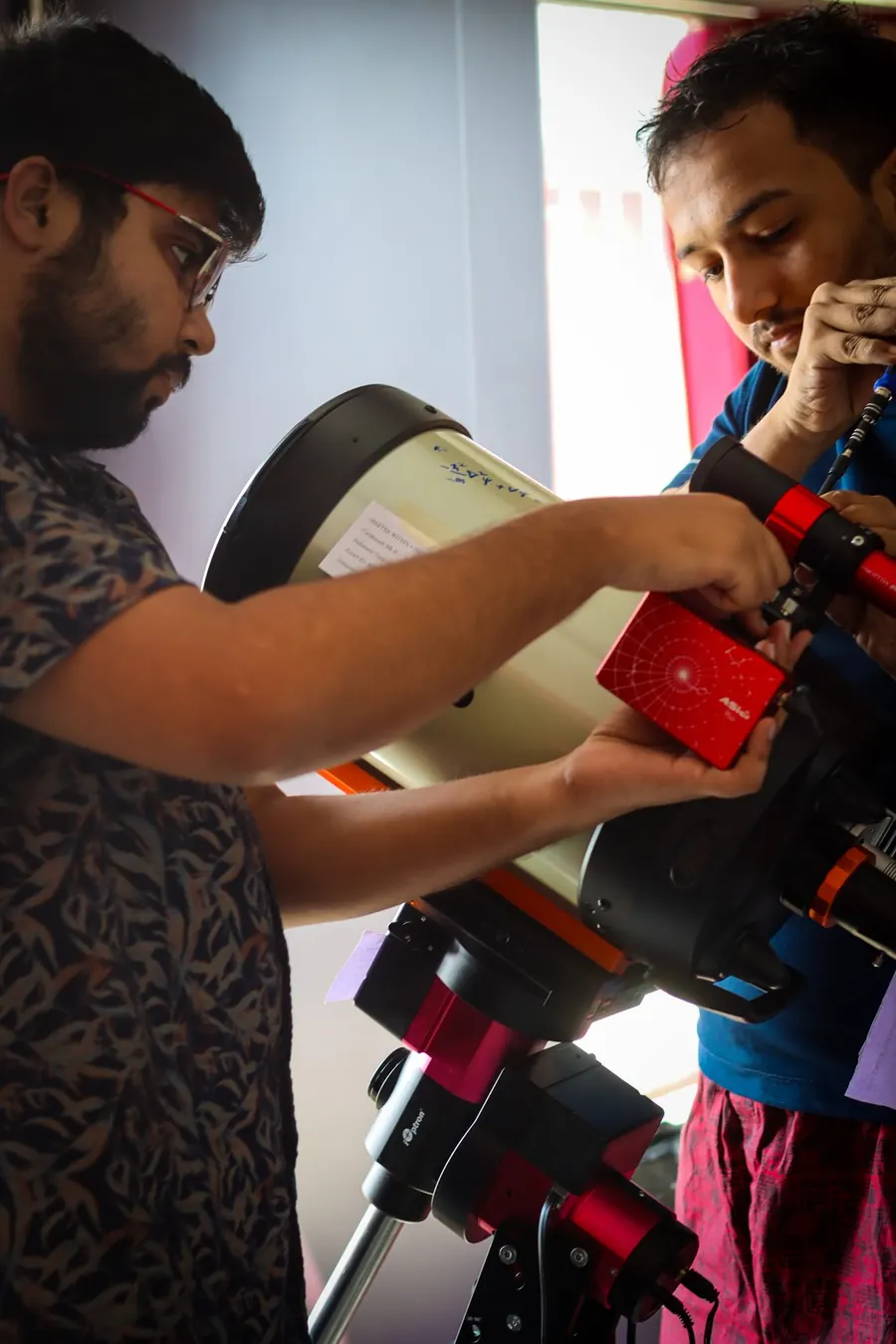 Two people fitting a red controller box and a guide scope to the back of the telescope tube.