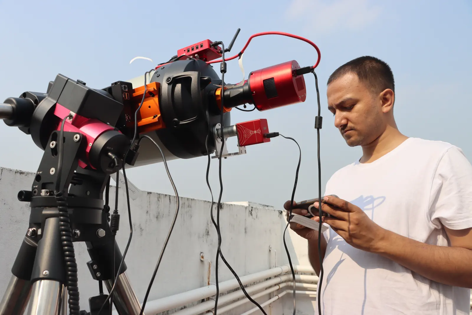 The Purākaash telescope on a rooftop in daylight, its cameras and cables attached, with Souvik Ghosh beside it holding the hand controller.